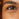A detailed close-up of a person's left eye area, highlighting the natural signs of aging. Visible crow's feet and fine lines crinkle outward from the corner of the eye and underneath the lower eyelid against a warm-toned complexion.