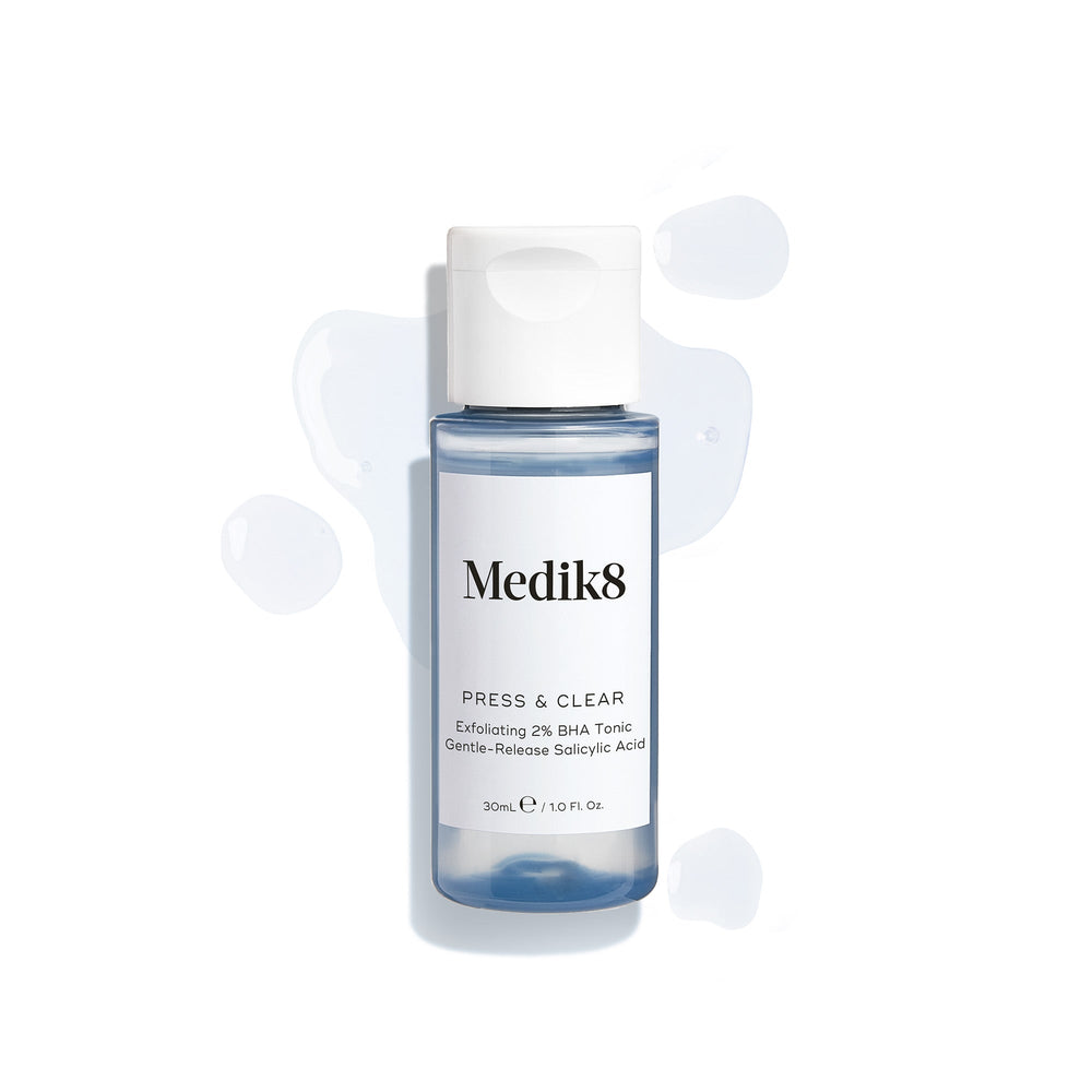 A travel-size bottle (30ml) of Medik8 Press & Clear tonic with a white flip-top cap, surrounded by clear blue droplets on a white surface. Text identifies it as an  Exfoliating 2% BHA Tonic. 