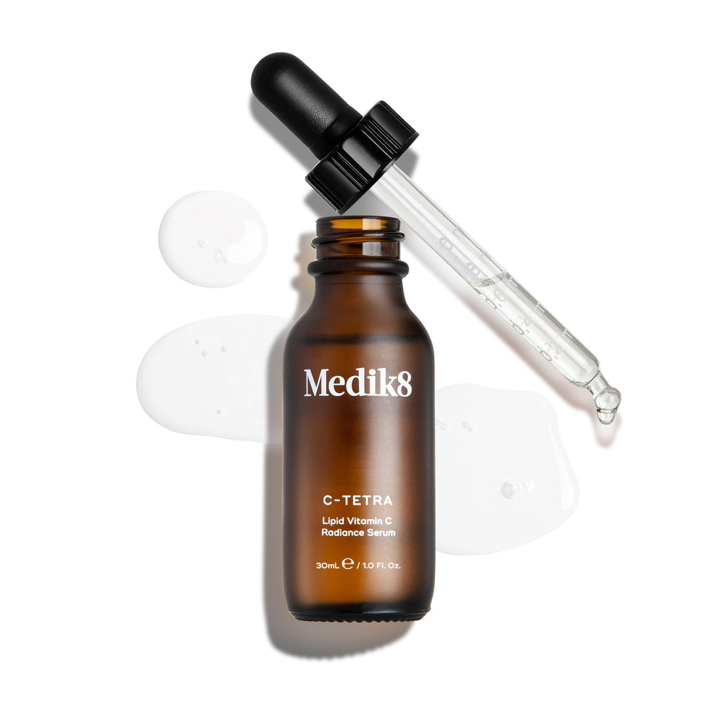 An overhead "packshot" of a 30ml bottle of Medik8 C-Tetra Lipid Vitamin C Radiance Serum. The amber glass bottle is uncapped and stands centered, with its black dropper lid resting diagonally behind it. A clear, liquid serum is artistically splashed and pooled on the clean white background around the bottle, with a few droplets visible on the glass pipette. The lighting is bright, casting soft shadows to the left and creating a clinical yet elegant aesthetic.