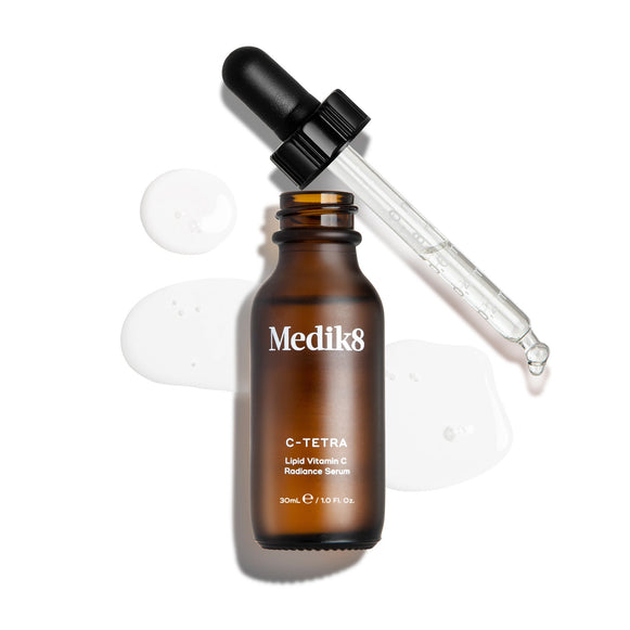 An overhead "packshot" of a 30ml bottle of Medik8 C-Tetra Lipid Vitamin C Radiance Serum. The amber glass bottle is uncapped and stands centered, with its black dropper lid resting diagonally behind it. A clear, liquid serum is artistically splashed and pooled on the clean white background around the bottle, with a few droplets visible on the glass pipette. The lighting is bright, casting soft shadows to the left and creating a clinical yet elegant aesthetic.-16
