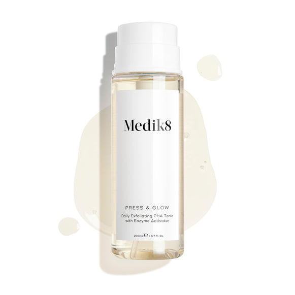 Medik8 Press & Glow tonic bottle stands against a clean white background with translucent liquid droplets and a puddle, highlighting its watery texture. Text identifies the product as a 200mL  Daily Exfoliating PHA Tonic with Enzyme Activator. -13