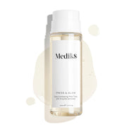 A 200mL clear refill bottle of Medik8 Press & Glow exfoliating tonic featuring a silver screw cap. Set against a white background, the bottle sits amidst a puddle and droplets of the pale yellow tonic, showcasing its liquid texture. The label text reads "Daily Exfoliating PHA Tonic with Enzyme Activator