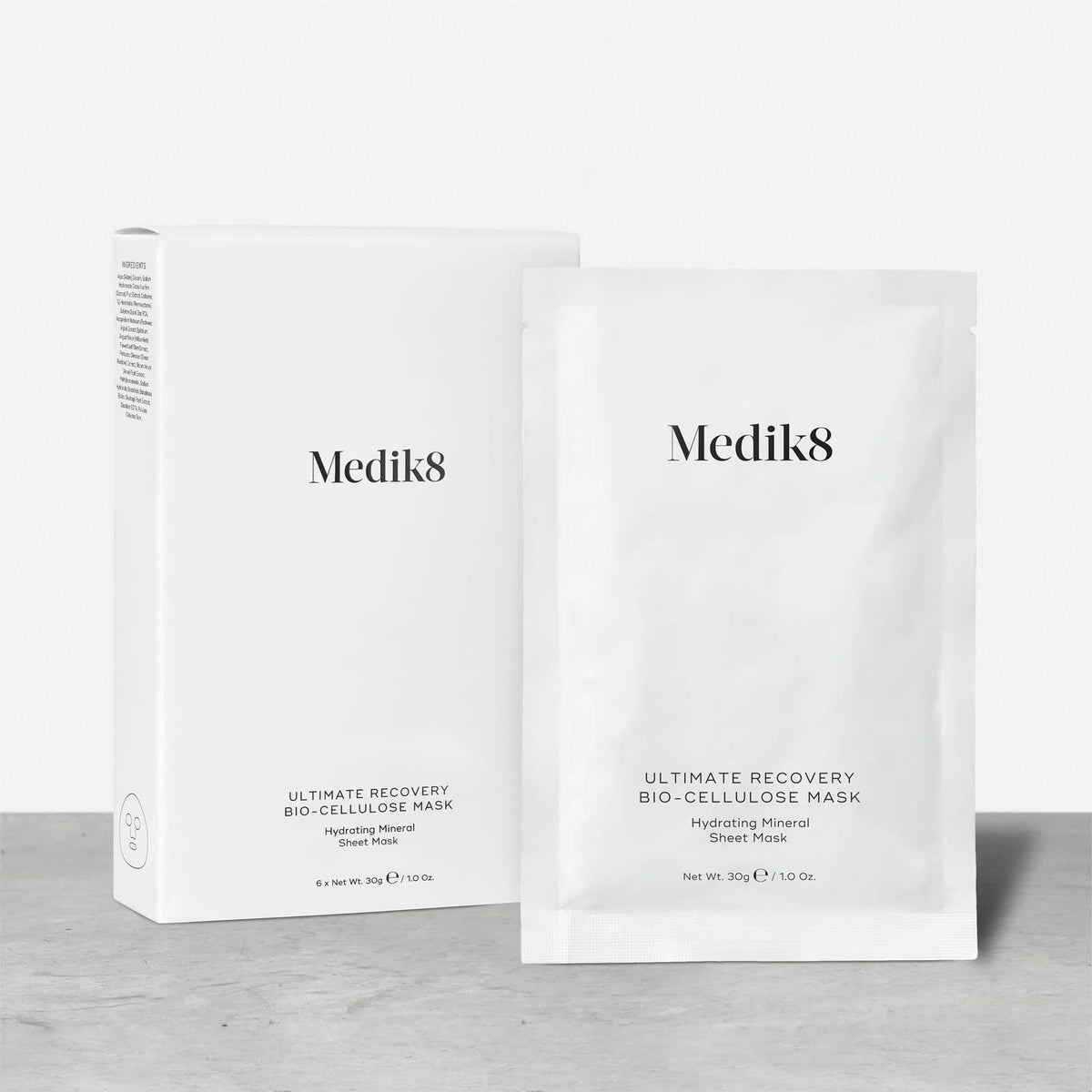 A white cardboard box of Medik8 Ultimate Recovery Bio-Cellulose Mask standing next to a single white sachet on a textured grey surface against a clean white background. Text indicates the box contains 6 sheet masks.