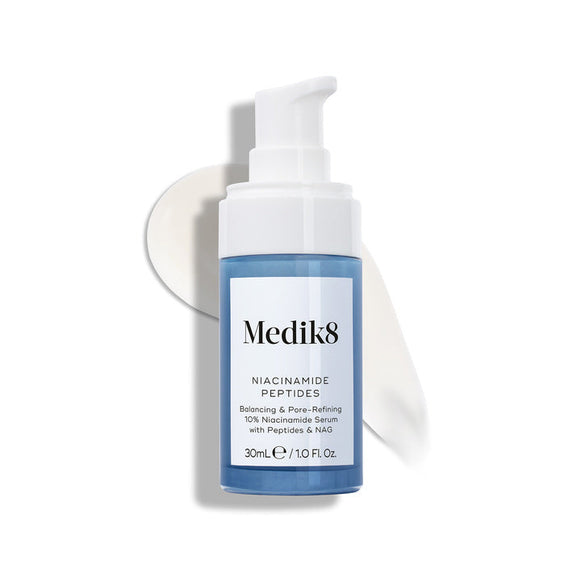 Medik8 Niacinamide Peptides serum bottle with a white pump top, resting on a white surface with a smear of white serum behind it.-47