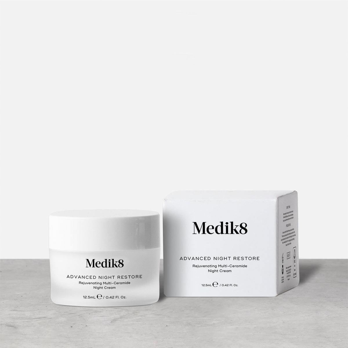 A travel-size jar (12.5ml) of Medik8 Advanced Night Restore cream displayed closed next to its box on a textured grey surface. Text identifies it as a  Rejuvenating Multi-Ceramide Night Cream. 