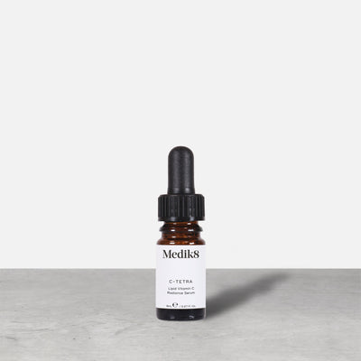 A travel-sized 8mL amber glass bottle of Medik8 C-Tetra lipid vitamin C radiance serum standing next to its white cardboard carton. The products are displayed on a textured grey surface against a clean white background