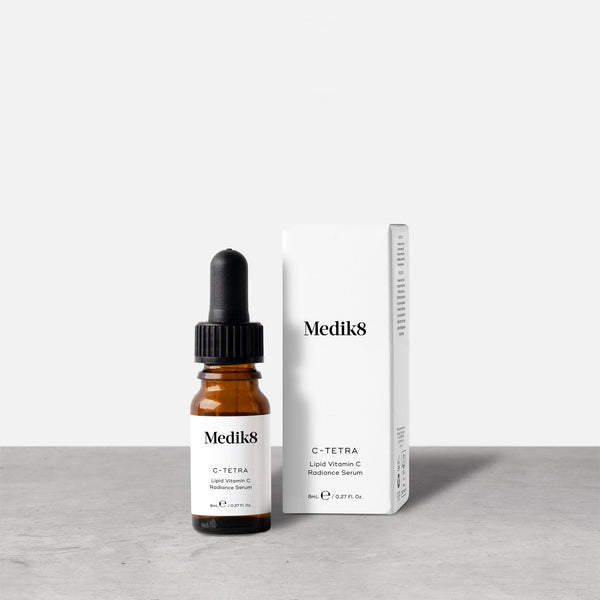 A travel-sized 8mL amber glass bottle of Medik8 C-Tetra lipid vitamin C radiance serum standing next to its white cardboard carton. The products are displayed on a textured grey surface against a clean white background.