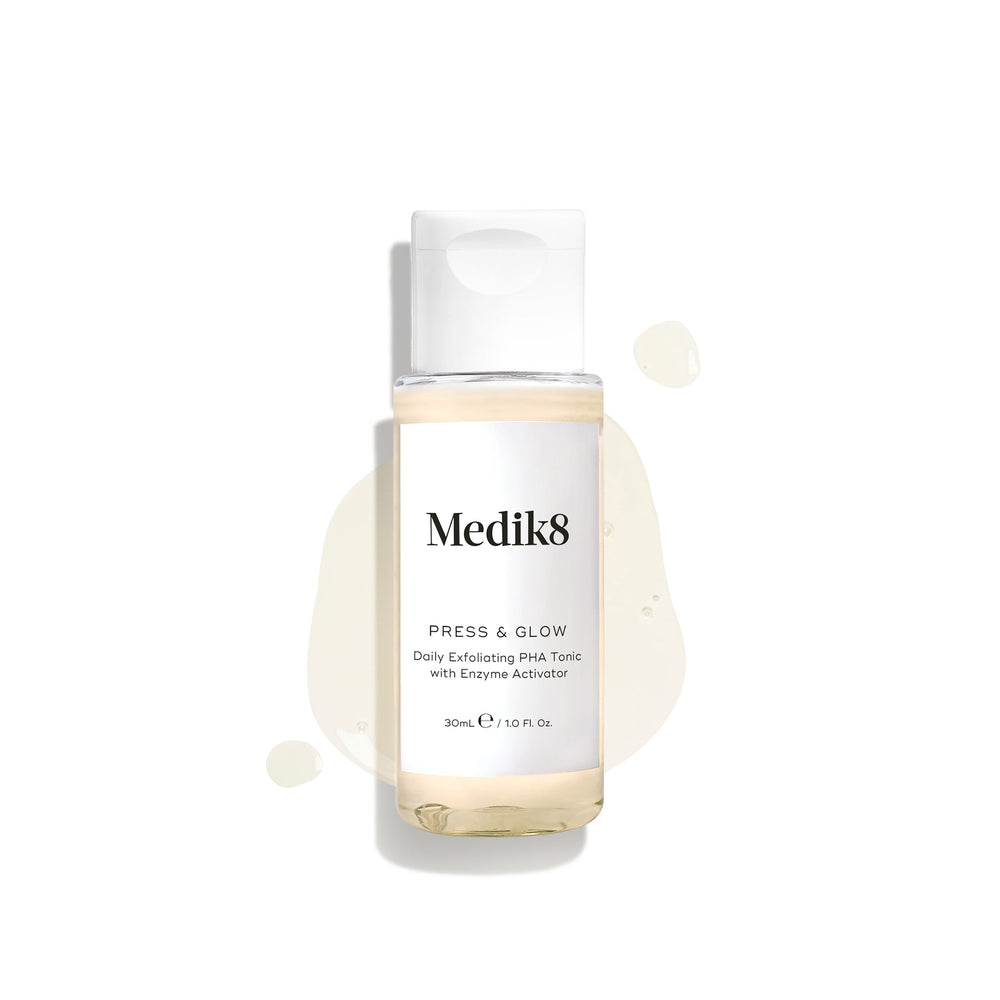 A 30mL travel-sized clear bottle of Medik8 Press & Glow tonic with a white cap, displayed on a clean white background. The bottle rests on a puddle of the pale yellow liquid with scattered droplets around it, highlighting its lightweight texture. Text on the label reads "Daily Exfoliating PHA Tonic with Enzyme Activator