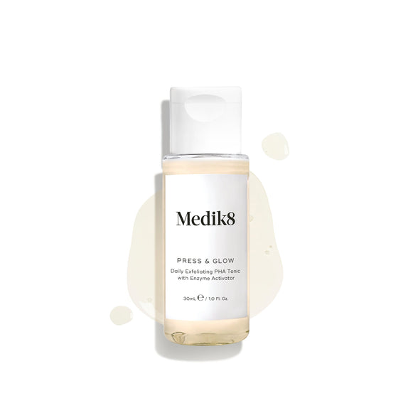 A 30mL travel-sized clear bottle of Medik8 Press & Glow tonic with a white cap, displayed on a clean white background. The bottle rests on a puddle of the pale yellow liquid with scattered droplets around it, highlighting its lightweight texture. Text on the label reads ""Daily Exfoliating PHA Tonic with Enzyme Activator.-4