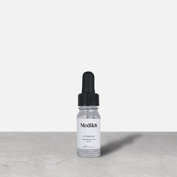 Medik8 Hydr8 B5 travel-sized serum bottle stand on a grey textured surface against a neutral background. Text on the 8mL bottle identifies it as a "Liquid Rehydration Serum.