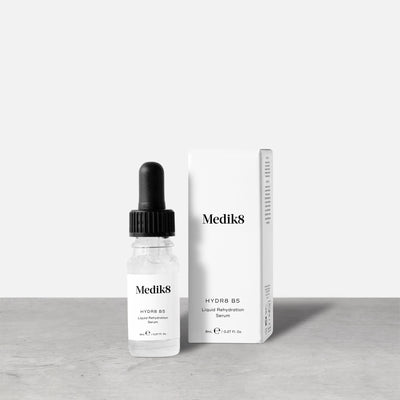 Medik8 Hydr8 B5 travel-sized serum bottle stands next to its matching white product carton on a grey textured surface against a neutral background. Text on the 8mL bottle identifies it as a "Liquid Rehydration Serum.