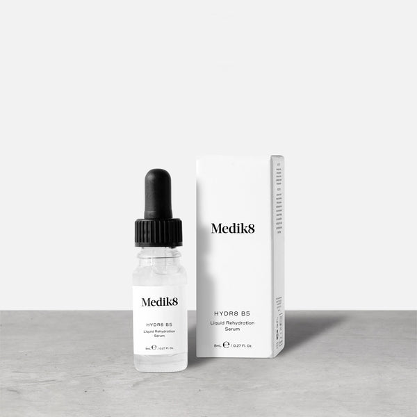 Medik8 Hydr8 B5 travel-sized serum bottle stands next to its matching white product carton on a grey textured surface against a neutral background. Text on the 8mL bottle identifies it as a "Liquid Rehydration Serum.