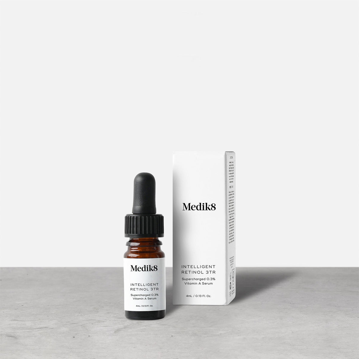 Medik8 Intelligent Retinol 3TR serum bottle stands next to its matching white product carton on a grey textured surface against a neutral background. Text on the 4mL bottle identifies it as a  Supercharged 0.3% Vitamin A Serum. 