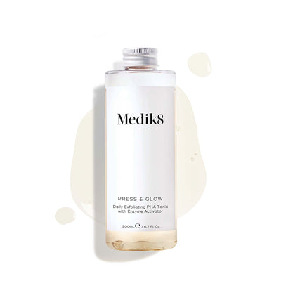 A 200mL clear refill bottle of Medik8 Press & Glow exfoliating tonic featuring a silver screw cap. Set against a white background, the bottle sits amidst a puddle and droplets of the pale yellow tonic, showcasing its liquid texture. The label text reads ""Daily Exfoliating PHA Tonic with Enzyme Activator."