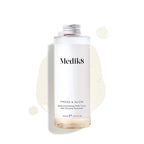 A 200mL clear refill bottle of Medik8 Press & Glow exfoliating tonic featuring a silver screw cap. Set against a white background, the bottle sits amidst a puddle and droplets of the pale yellow tonic, showcasing its liquid texture. The label text reads ""Daily Exfoliating PHA Tonic with Enzyme Activator."""